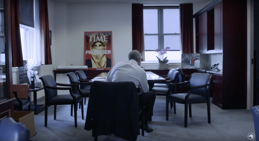 a man sits at a conference table with his back to the camera. A poster-sized Time magazine cover hangs on wall in the background