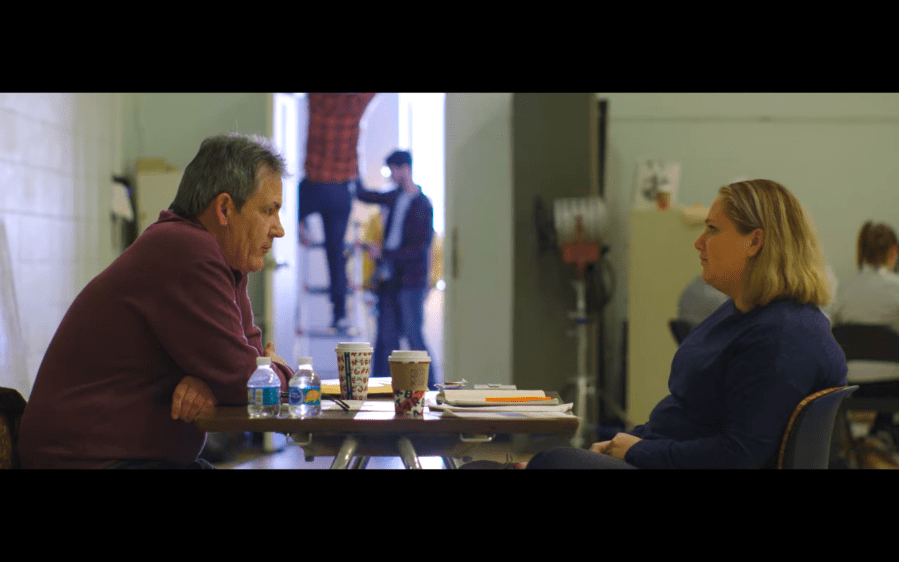 A middle-aged man sits at a craft services table with a female drama therapist while two men set up lights on set in the background
