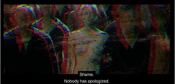 Blurry image of topless woman, surrounded by men, gazing straight at the camera