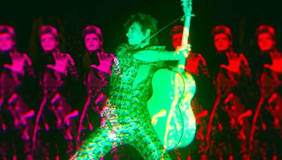 A green David Bowie in the foreground while 6 identical copies of David Bowie in pink and red look on from the background