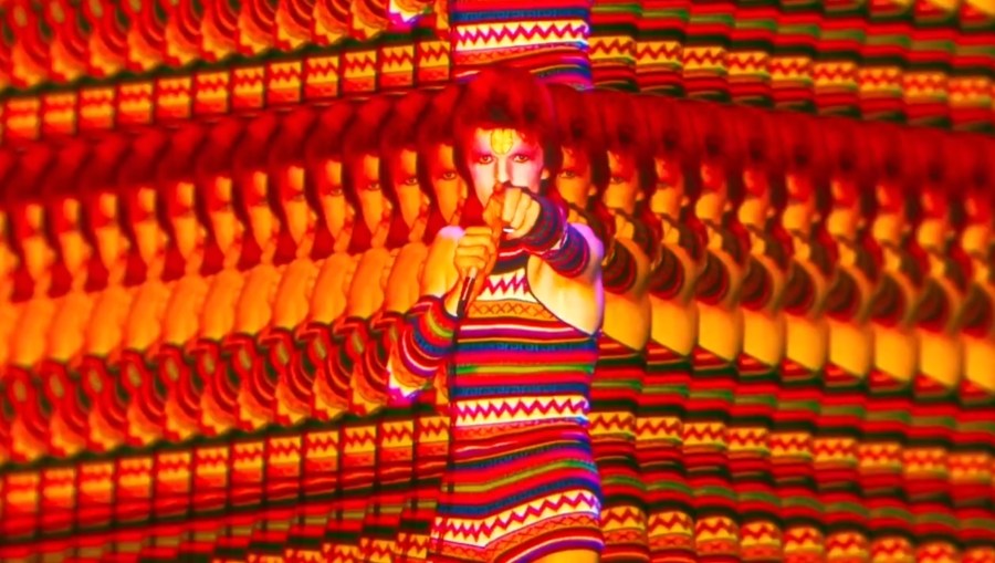 A singer, performing, pointing to the audience, with a mirror image effect surrounding him