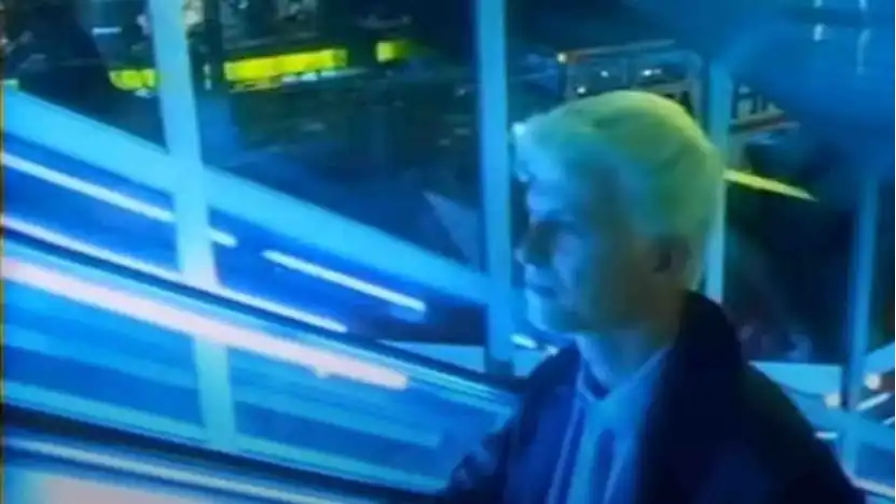 A blonde haired man rides a blue neon-drenched escalator