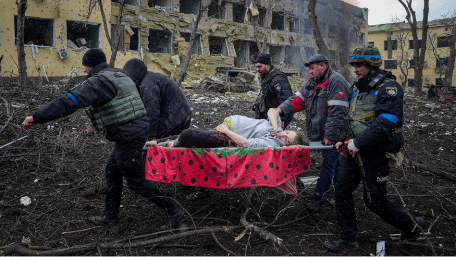 A preganant woman on a gurney being carried by 5 men through a bombed out hospital courtyard
