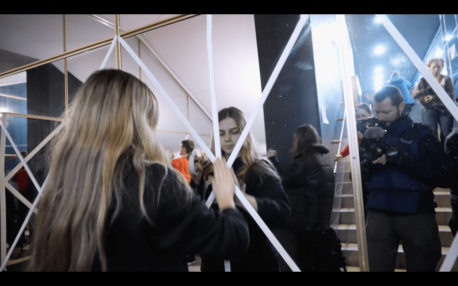 A young woman with long blond hair puts protective tape on a wall mirror in which we see the reflection of the camerman as he looks on