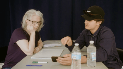 a woman stares at a cell phone while a man points at the image on it